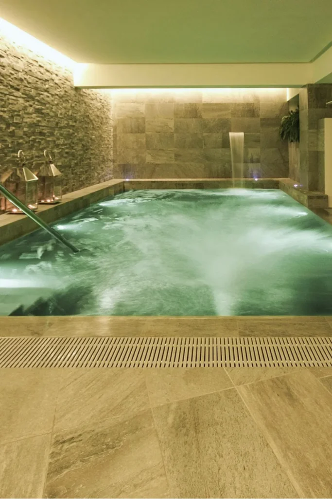 A minimalist, modern spa bathroom featuring a clean-lined soaking tub set against a textured wood-slat wall. The room is flooded with natural light from a large window, which reflects off the water's surface to create "dancing reflections" on the warm-toned walls. The design showcases the use of water as a visual anchor, complemented by organic textures like a woven stool and neutral stone flooring to create a silent, luxurious retreat.