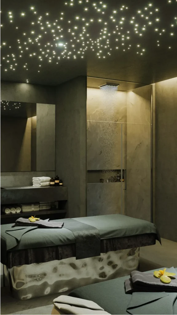 A dark, ultra-modern luxury treatment room featuring a fiber-optic "starry sky" ceiling that casts a soft glow over a stone-base massage table. The room emphasizes hidden storage and low profiles, with a seamless stone vanity holding only a neat stack of white towels. In the background, a glass-enclosed rain shower with integrated warm lighting serves as a focal point. The design follows the "hide everything" principle, removing all clutter and sharp corners to create a grounded, serene environment for healing.