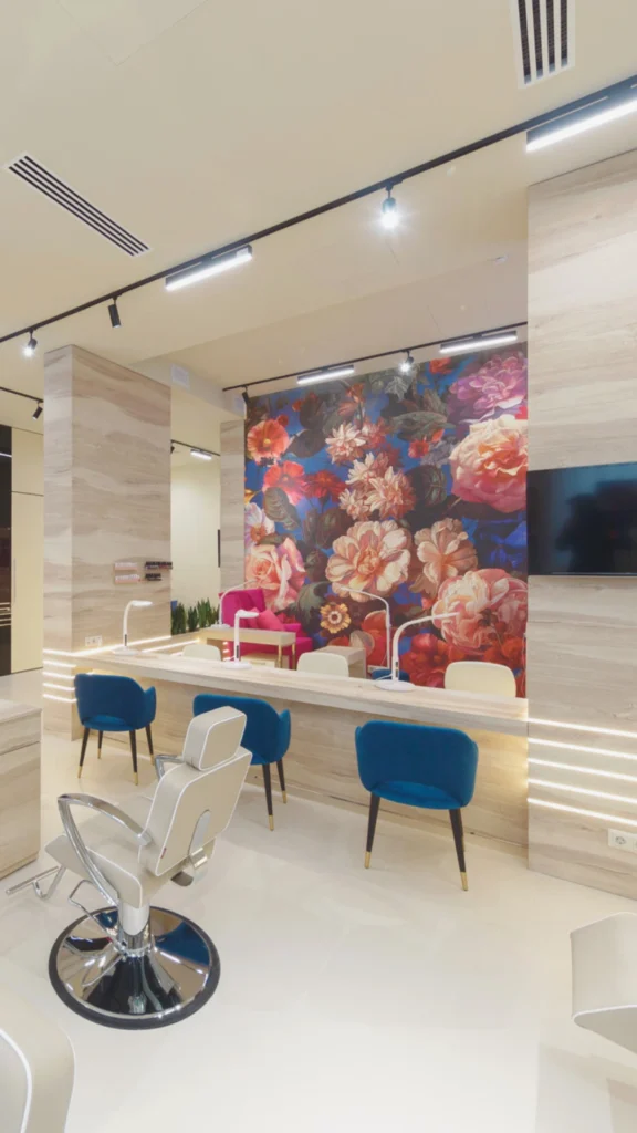 A vibrant, upscale beauty salon featuring a long, light wood manicure or service bar lined with elegant royal blue velvet chairs with tapered legs. In the foreground, a premium beige leather styling chair with high-shine chrome accents sits on a pristine, polished white floor. The standout feature is a magnificent, large-scale accent wall covered in a rich floral mural showcasing oversized pink, red, and orange blossoms against a deep blue background. The space is framed by textured light wood pillars with integrated, horizontal LED strip lighting, all illuminated by a modern black track lighting system on the ceiling.