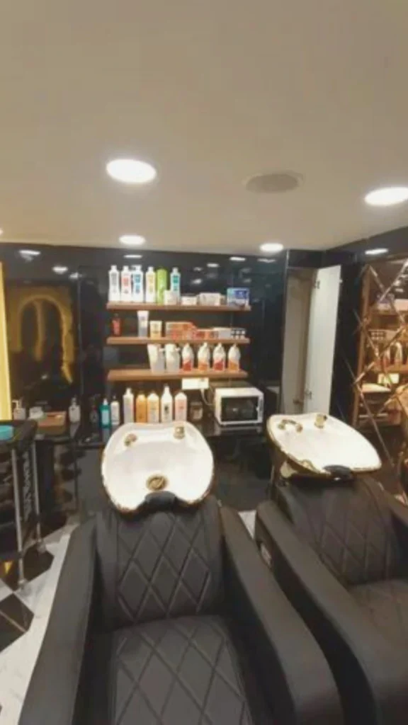 A dedicated, premium shampoo and rinse station inside a luxury hair salon designed for quiet relaxation. Two plush, black diamond-tufted leather reclining chairs are paired with deep white ceramic washbasins accented with elegant gold fixtures. The space features a sleek black gloss back wall lined with floating wooden shelves that house neatly organized salon products and a small towel warmer. The area is dimly lit with recessed warm ceiling spotlights, creating a serene, private mini-escape for clients.