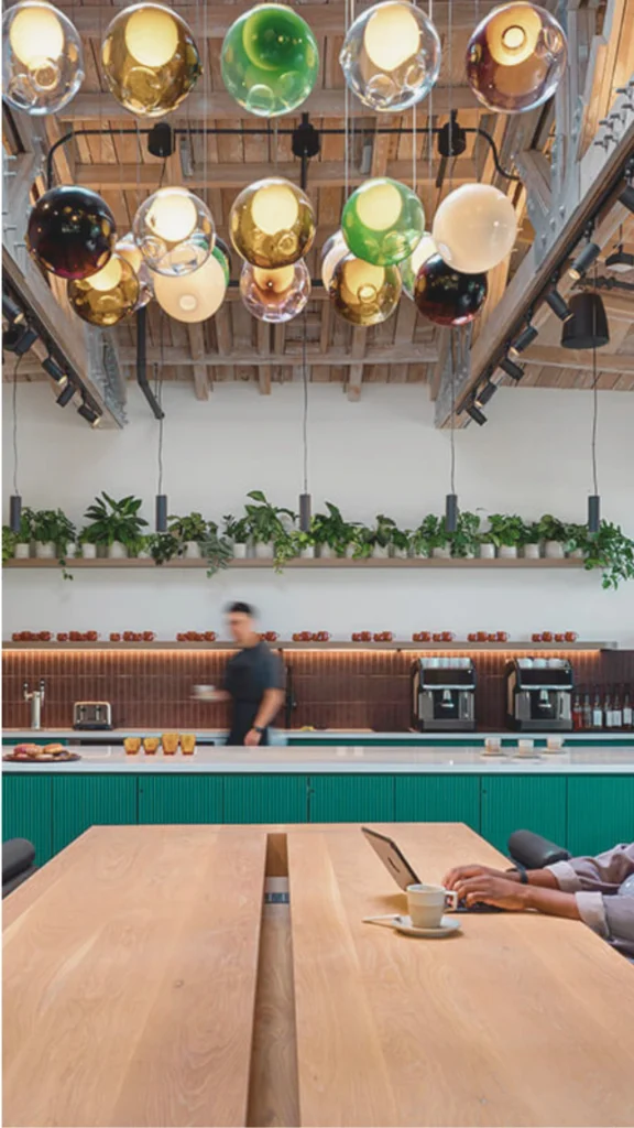 A spacious, modern office lounge or cafe featuring a high, vaulted rustic wooden plank ceiling with exposed timber beams and skylights. Hanging centrally is a striking cluster of colorful, glass-globe pendant lights in shades of amber, emerald green, clear, and white. In the foreground, a man sits at a long, light wood communal table working on a laptop next to a coffee cup. In the background, a sleek, long service counter with a rich teal green base and white countertop houses coffee machines, while a woman talks to a staff member. The back wall is decorated with a long floating shelf crowded with small potted green plants, and a large window on the right lets in abundant natural daylight.