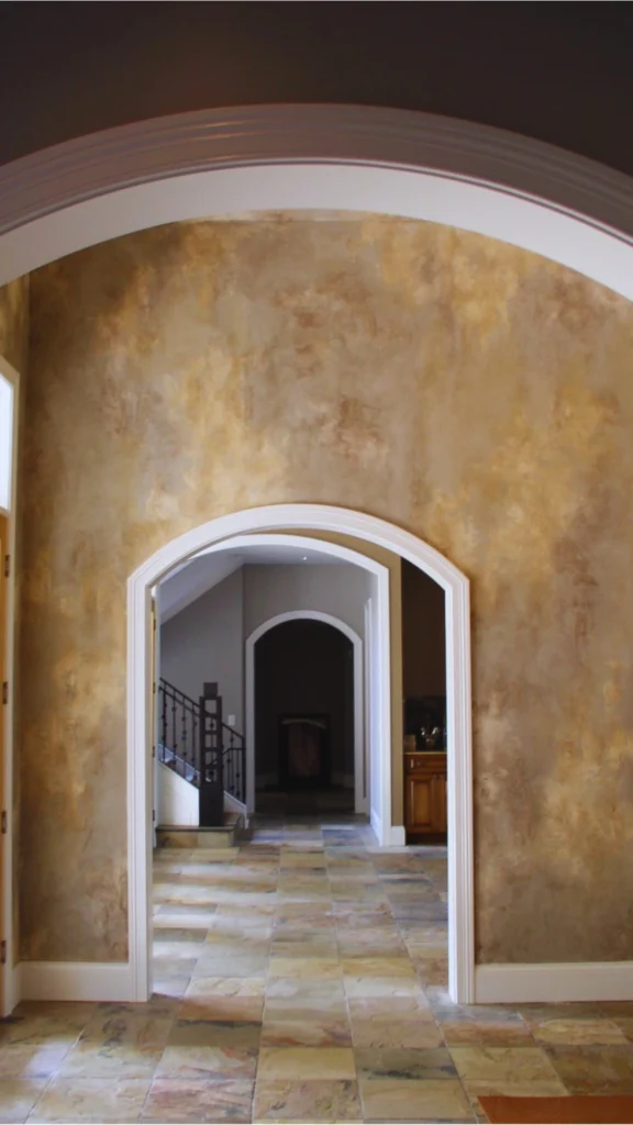 A long interior hallway showcasing textured luxury wall design with sequential arched openings. The walls are finished in a warm, hand-troweled Venetian plaster featuring a mottled blend of beige, gold, and soft brown tones that catch the light. A white architectural archway frames the view into the hallway, leading the eye through a second matching archway toward a dimmer background area with a staircase and a dark wood cabinet. The floor is laid with square natural slate tiles in variegated earthy shades of gray, tan, and rust.