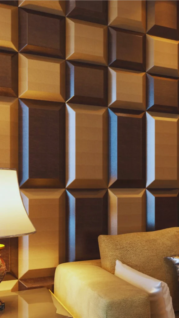 A close-up shot showcasing a striking 3D upholstered accent wall designed with a bold geometric pattern. The wall is covered in dimensional, rectangular and square padded tiles arranged in a staggered checkerboard layout, featuring a mix of rich chocolate brown leather and textured golden-bronze fabric. The beveled edges of the panels create crisp shadow lines and a deep sense of touchable texture. In the foreground, the warm yellow glow of a table lamp illuminates the corner of a plush, textured beige sofa styled with accent pillows, highlighting the room's luxurious and acoustically padded design.