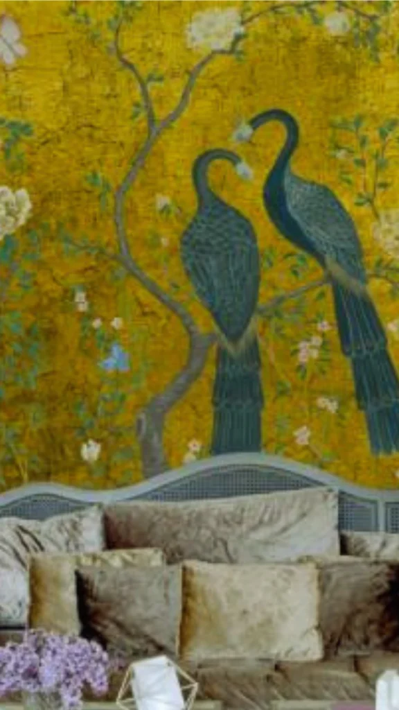 A close-up view of an upscale living space showcasing an exquisite Chinoiserie-style custom wallpaper mural. The wallpaper features a highly textured, shimmering golden background decorated with slender tree branches, white blossoms, and two large, detailed peacocks with deep teal and blue plumage perched side by side. Below the feature wall sits a gray cane-back vintage sofa generously layered with plush velvet throw pillows in shades of metallic gold, bronze, and charcoal gray. In the bottom left corner, a bouquet of purple lilac flowers in a glass vase rests on a coffee table, complementing the room’s artful and luxurious atmosphere.
