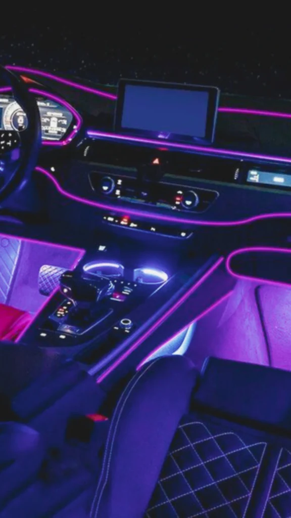A futuristic luxury vehicle interior showcasing vibrant, customized ambient LED lighting at night. Neon magenta and purple light strips trace the sleek contours of the dashboard, instrument cluster, and center console, emphasizing the car's high-tech architecture. Soft purple illumination glows from the footwells and the integrated cup holders on the central console. In the foreground, a dark premium leather sport seat is visible, featuring detailed white diamond-quilted stitching that catches the ambient colored light.