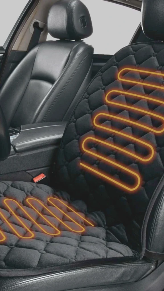 A close-up demonstration of an active vehicle seat heating system installed inside a premium car cabin. The image showcases a black plush, diamond-tufted and quilted seat cover draped securely over a dark leather car seat. Superimposed over the seat cushion and backrest are glowing, orange neon zigzag lines that trace a heating element graphic, visually representing warm, temperature-controlled thermal zones. The surrounding car interior, including the sleek black center console and leather passenger seat, remains subtly in focus in the background.
