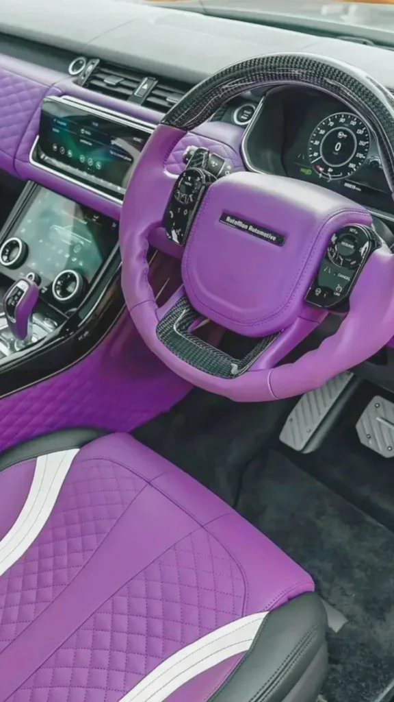 A view looking down into the highly customized, vibrant purple driver's side cockpit of a luxury vehicle. The multi-functional sport steering wheel is wrapped in premium bright purple leather with a glossy carbon fiber top and bottom rim. The dashboard, center console trim, and upper door inserts are wrapped in a matching purple leather, while the lower sections feature a diamond-quilted purple leather texture. In the foreground, a black leather sport seat is detailed with a purple diamond-stitched center and sweeping white accents. The digital infotainment screen and instrument cluster are powered on, adding to the high-tech, custom aesthetic.