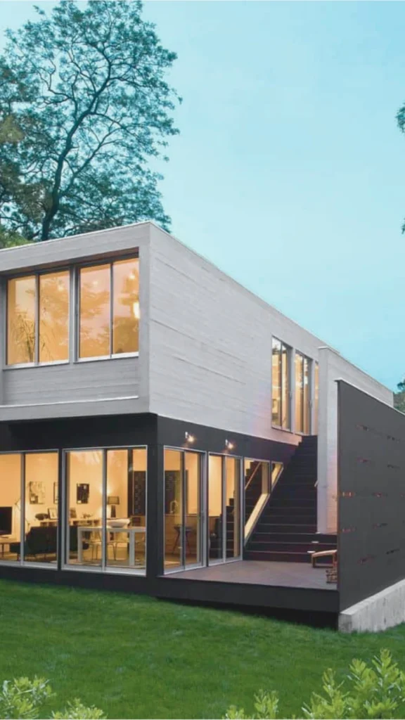 A striking two-story luxury container home featuring a clean, minimalist architectural layout. The structure blends industrial elements with modern design, showcasing a ground level constructed from deep matte-black steel frame sections encased in seamless, expansive floor-to-ceiling glass sliding doors. The bright, warmly lit interior living spaces are fully visible from the lush green lawn outside. The upper level consists of a contrasting light gray, textured concrete-style container volume with wide glass windows. An exterior black wooden staircase ascends along a sleek dark privacy screen wall, connecting the ground patio deck to the upper living quarters under a clear blue evening sky.