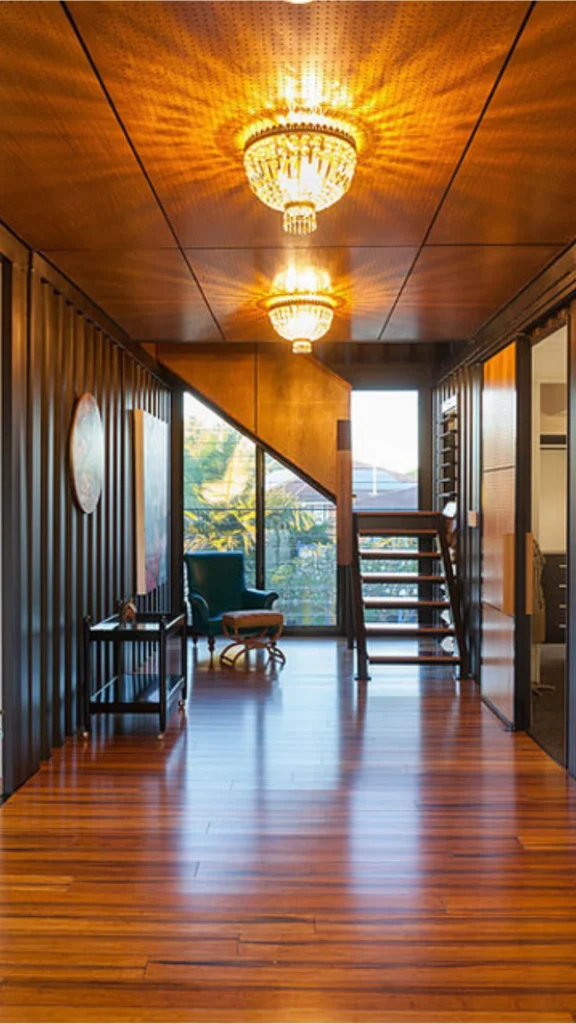 A striking close-up of a modern container home's exterior corner, illustrating high-end container home design through contrasting textures. The building’s industrial, deep matte-black corrugated steel paneling is paired seamlessly with a vertical slat wall made of warm, light cedar siding. A clean, minimalist black metal corner trim creates a sharp boundary between the rustic wood and the dark steel. Lush green tree leaves in the soft-focus background catch the gentle sunlight, adding a natural touch to the architectural details.