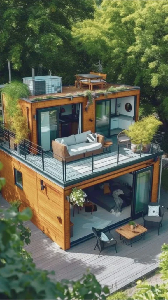 A high-angle view of a luxurious, multi-level shipping container home beautifully integrated into a lush, green landscape. The house features rich, warm honey-toned wooden siding juxtaposed with black metal frames and large glass sliding doors that completely open up the living spaces. On the second-floor balcony deck, an outdoor seating area is arranged with a comfortable wicker sofa and plants. The upper roof is utilized as a green terrace featuring a small wooden dining set, trailing plants, and low-profile utility systems. Below, the ground level opens onto a light gray wood-plank patio adorned with stylish outdoor armchairs and a small table.