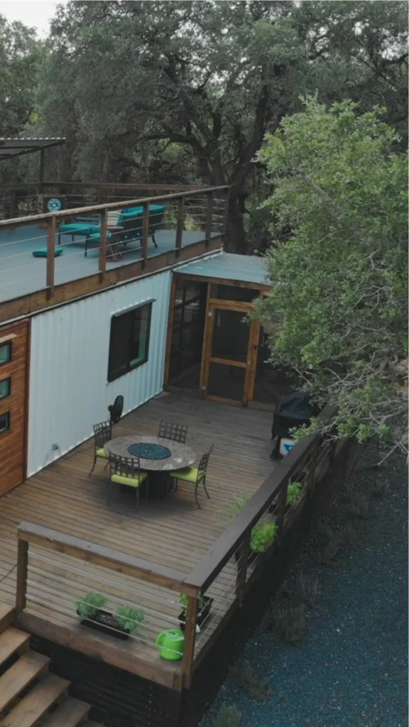A high-angle architectural shot of a modern multi-level shipping container home featuring expansive outdoor living spaces. The lower shipping container volume is finished in clean white siding with dark-framed windows and wood accents, opening onto a spacious lower wooden deck. The deck is arranged with an outdoor dining table, chairs with vibrant lime-green cushions, and low planters along the wooden railing. A wooden staircase leads to the flat roof of the lower container, which has been transformed into an upper lounge deck complete with bright teal patio recliners. Mature canopy trees surround the property, creating a natural and private setting.