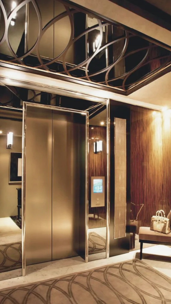 A striking vertical shot of a premium elevator interior design showcasing high-end materials and sophisticated architecture. The focal point features sleek, champagne-gold brushed metal sliding doors framed by highly reflective mirrored side panels that stretch floor-to-ceiling. Above, the polished dark ceiling features an intricate, geometric metal overlay of overlapping curved circles, which beautifully reflects the warm, ambient lighting. The adjacent accent walls are lined with a rich, textured dark wood or fabric finish. On the right, a luxurious beige patterned area rug mirrors the circular motif of the ceiling, lying near a plush waiting chair holding a luxury handbag.