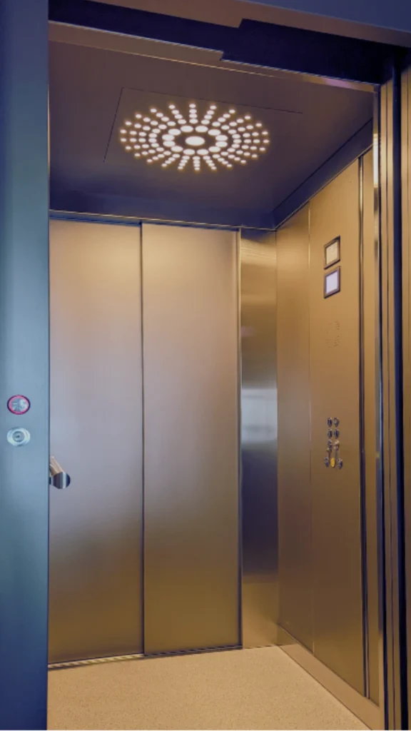 A view looking inside an open elevator cab, highlighting an elegant lift interior design centered on unique decorative lighting. The cabin is constructed from sleek, brushed champagne-gold or bronze metal panels, featuring a smooth sliding back door and an adjacent control panel with polished buttons. Embedded in the flat, dark ceiling is the main design feature: a square light fixture radiating a circular, starburst pattern of warm yellow pinholes that glow outward from a central point. The soft lighting reflects gently off the metallic walls, casting a warm ambiance over a light-colored, subtly textured floor.
