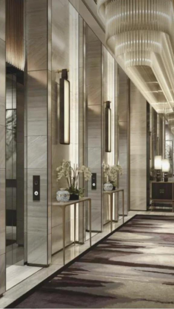 A long shot of an opulent hotel or residential lobby corridor highlighting premium elevator interior and hallway integration. The wall features a sequence of elevator bays clad in polished, light grey veined marble panels interspersed with tall vertical mirrors. Slender bronze console tables are placed between the elevator doors, each decorated with white orchids and patterned ceramic vases. Sophisticated lighting layers the space, featuring long, minimalist black wall sconces, soft warm backlighting behind the marble pillars, and a grand, tiered crystal chandelier stretching across the ceiling. On the floor, a luxurious, large-format area rug with deep burgundy and cream abstract brushstroke patterns creates a rich contrast down the length of the hallway.