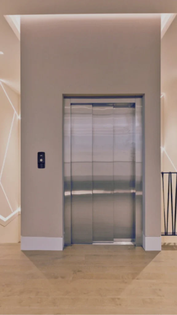 A straight-on view of a residential or boutique hotel elevator bay set in a minimalist, modern hallway. The elevator features brushed stainless-steel sliding doors inset within a smooth, beige-colored column wall. To the left of the door, a sleek, black digital smart interface panel is mounted for touchless controls or calls. The surrounding walls are illuminated by geometric linear light fixtures that trace glowing abstract patterns in the background. The floor is made of light-colored hardwood planks, bathed in warm, soft overhead lighting that complements the clean, contemporary architecture.