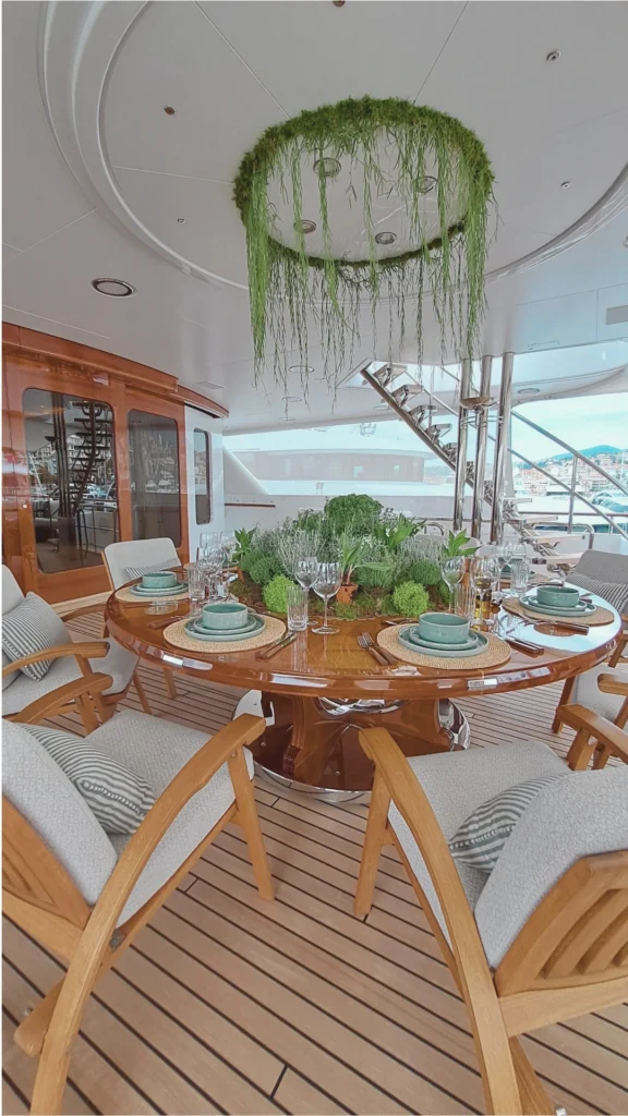 A vertical view of an open-air luxury boat dining deck from the file, highlighting fine wood finishes and organic design elements. A large, round dining table crafted from high-gloss, polished wood features a central decorative centerpiece filled with lush green mosses, herbs, and potted plants. The table is elegantly set with woven circular placemats, light teal ceramic dinnerware, fine cutlery, and crystal wine glasses. Surrounding the table are matching wooden armchairs fitted with textured grey cushions and striped pillows, arranged over a classic striped teak wood deck floor. Directly above the table, a unique, circular ring fixture hangs from the white paneled ceiling, decorated with trailing, vine-like green plants. In the background, a glossy wooden bulkhead door, polished steel support pillars, and an outdoor staircase are visible, with a glimpse of a coastal landscape on the horizon.