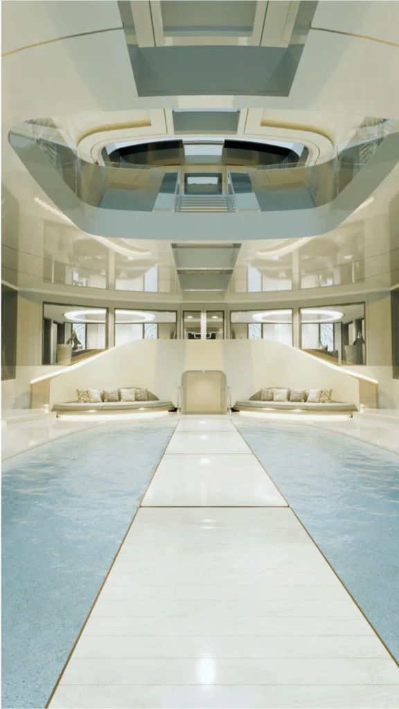 A highly futuristic, ultra-luxurious indoor pool deck and multi-level atrium inside a superyacht. A central white marble runway splits a glistening light-blue swimming pool into two halves. At the far end of the pool, a symmetrical white grand staircase curves upward, flanked by integrated curved bench seating with textured neutral throw pillows. The architecture above features dramatic, soaring cutouts looking into the upper decks, enclosed by sleek glass railings. The entire interior has a polished, reflective high-gloss finish with clean geometric lines, soft integrated LED accent lighting, and a neutral, cream-and-white color palette.