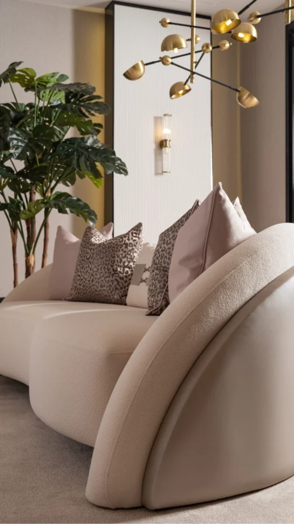 A close-up, angled view of a modern luxury sofa with a unique, sculptural curved back and a mix of smooth leather and textured fabric finishes. The sofa is styled with an intentional arrangement of plush pillows, including soft mauve tones and a leopard print pattern, demonstrating dynamic texture mixing. In the background, a large Monstera plant adds a touch of natural greenery, while a striking gold multi-arm chandelier and a minimalist wall sconce provide sophisticated ambient lighting. The scene emphasizes a clean, intentional aesthetic where "less is more," leaving breathing room to highlight the furniture's high-end design.