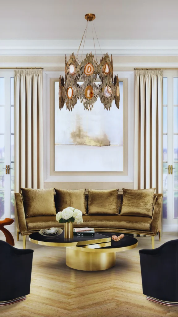 A symmetrical, upscale living room arrangement showcasing a "conversation zone" layout. A plush, gold velvet sofa is the centerpiece, set slightly away from the wall and framed by a large abstract painting and cream-colored drapes. In front of the sofa, a tiered black and gold circular coffee table is positioned with ample walking space, flanked by two dark accent chairs in the foreground. Overhead, a dramatic sculptural chandelier with gold and agate accents draws the eye upward, while the light wood herringbone floors and clean sightlines emphasize a curated, luxury living room aesthetic.