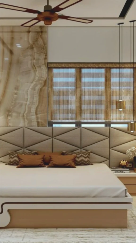 A detail-oriented view of a modern luxury bedroom showcasing a variety of rich textures and materials. The bed features an oversized headboard with a geometric, triangular quilted fabric pattern in a warm neutral tone. It is styled with several brown and chevron-patterned accent pillows. Behind the bed, the wall is anchored by a large, polished marble panel with swirling organic veins, paired with natural-fiber horizontal blinds. The scene is completed with a sleek white bed frame, a minimalist nightstand holding a gold sculptural vase, and slender cylindrical gold pendant lights, exemplifying an elegant bedroom design through layered tactile elements.