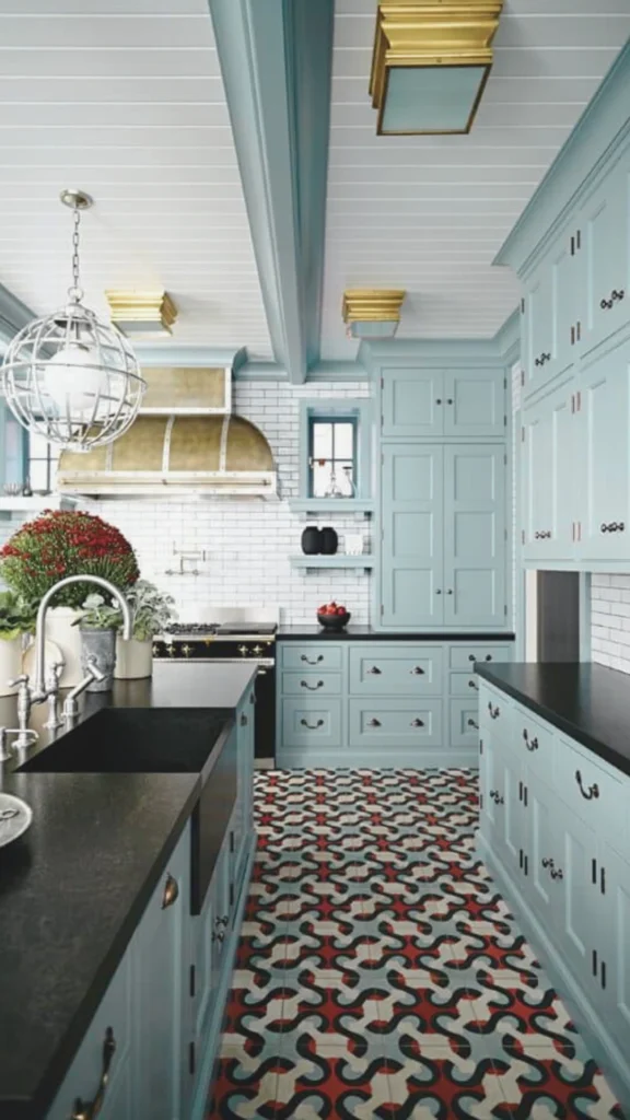 A beautifully lit, high-end kitchen displaying multiple layers of architectural and decorative lighting. The space features custom pale blue cabinetry contrasted by solid black countertops and a bold, patterned red, white, and blue tiled floor. Task lighting is provided by a large, sculptural silver globe cage pendant light hanging over the island's black farm sink, while warm accent lighting glows from under-cabinet LED strips against a white subway tile backsplash. The look is completed by unique brass flush-mount ceiling fixtures and a large brushed brass range hood, creating a bright yet warm, conversational atmosphere.