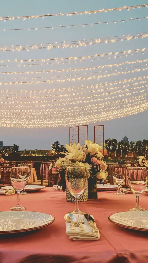 A grand banquet hall beautifully transformed by sophisticated event lighting. Dozens of warm, glowing crystal chandeliers hang in rows from a high ceiling, casting a dramatic, romantic light over the room. The long reception tables are meticulously set with white linens, tall floral centerpieces, and glowing candles that add layers of intimate illumination. The perimeter of the room features soft up-lighting against elegant architectural wall panels, highlighting a breathtaking and luxurious atmosphere designed for high-end celebrations.