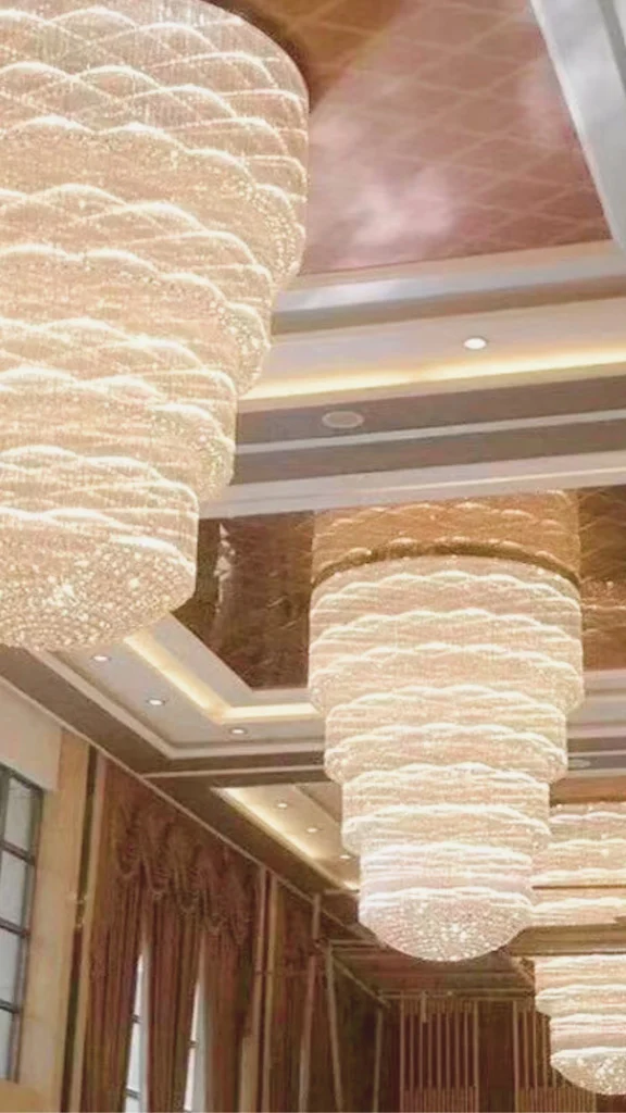 A close-up, upward perspective of a luxury banquet hall ceiling featuring a row of massive, multi-tiered crystal chandeliers. Each showstopping chandelier features a magnificent cascading design made up of thousands of shimmering crystal droplets that create a brilliant wave-like pattern. The chandeliers are set against a reflective, metallic copper-toned mirrored ceiling trim with embedded warm recessed lights, demonstrating how large-scale, high-end lighting can completely transform a grand venue space.