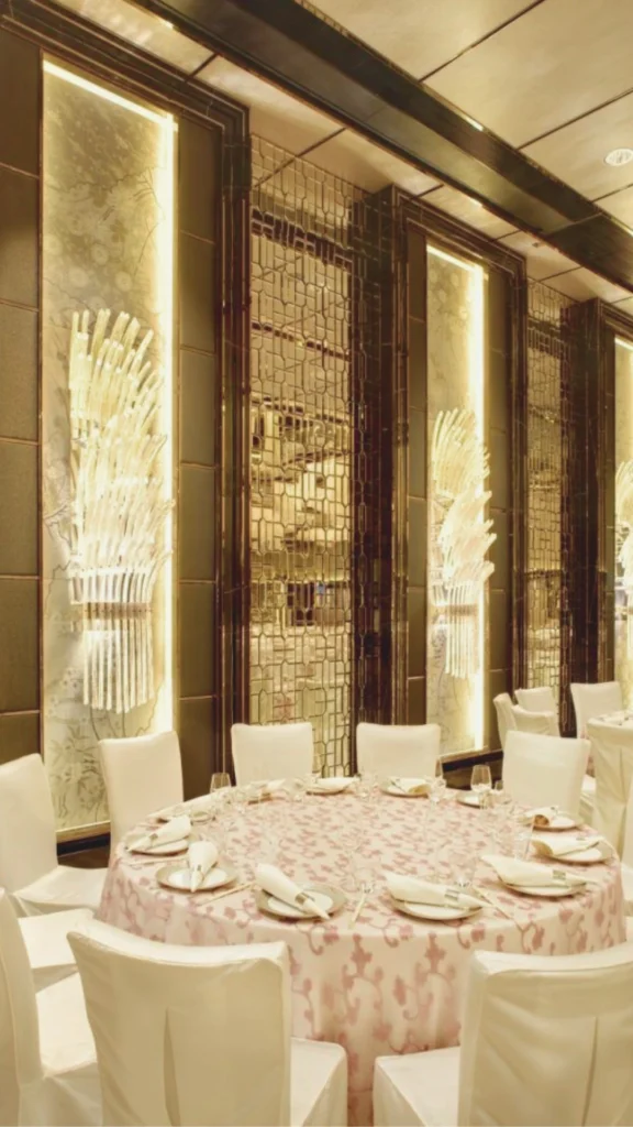 A grand, symmetrical banquet hall showcasing elegant wall decor and a layered lighting scheme. The center of the room features perfectly arranged round tables with white linens and dark wooden chairs, arranged over a patterned red and gold carpet. Hanging from the coffered ceiling are three massive, multi-tiered crystal chandeliers shaped like shallow bowls. In the background, the primary feature wall is adorned with a large, custom floral mural framed by rich wood paneling, perfectly positioned to capture the ambient warmth of the room and create an instant sense of luxury.