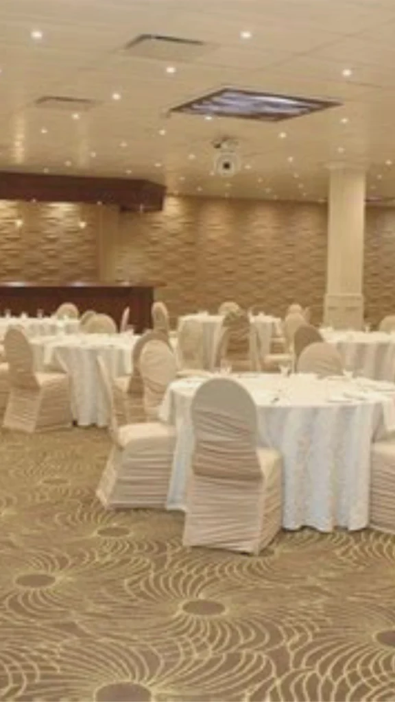 A striking luxury banquet hall showcasing the power of warm white lighting to create a rich and intimate atmosphere. The high ceiling is adorned with long, modern rectangular crystal chandeliers that cast a deep amber-toned glow over a dark, highly reflective glossy floor. In the center, a long reception table is styled with crisp white linens and tall, dramatic floral arrangements that catch the light perfectly. The stark contrast between the bright, warm gold light and the deep shadows exemplifies how programmable dimming levels can instantly transform a grand event space.