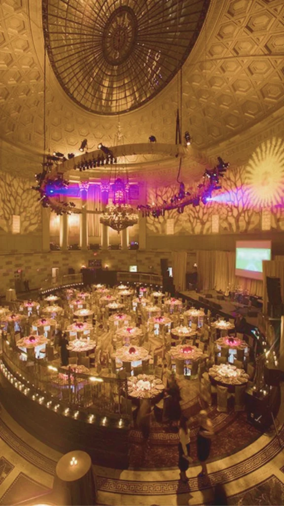 A breathtaking, high-angle panoramic view of an ultra-luxury, circular banquet hall showcasing dramatic lighting accents. An immense, intricate domed ceiling with a central glass skylight towers over the space, illuminated by an immersive gobo light projection casting an organic tree-branch shadow pattern across the sweeping walls. A large, circular professional lighting rig suspended from the ceiling projects vibrant purple and gold spotlights onto the grand event floor below. Dozens of round banquet tables are densely arranged and glow with warm, clustered centerpieces, while rows of tiny fairy lights trace the curved balcony perimeter, creating an unforgettable, star-lit atmosphere.