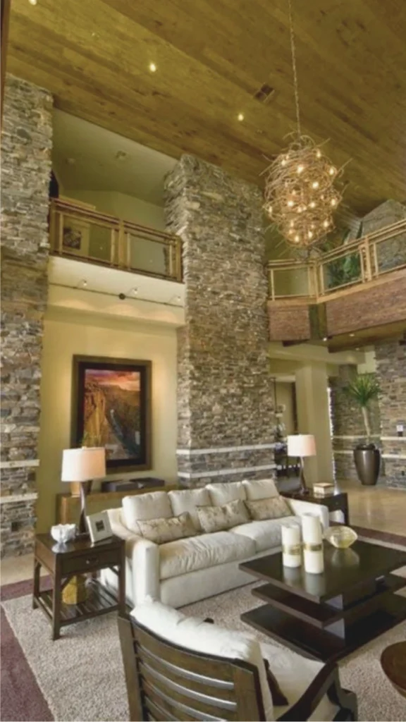 A grand, double-height luxury villa living room showcasing rustic yet upscale stone foundations and architecture. Two massive, floor-to-ceiling accent pillars constructed from textured, multi-toned stacked ledger stone frame the central seating area and support a high, warm wood-paneled ceiling. The space features an elegant cream-colored fabric sofa paired with a dark wood tiered coffee table, sitting beneath a large, sculptural branch-like orb chandelier. In the background, a second-story mezzanine balcony with wood and glass railings overlooks the expansive, stone-clad interior.