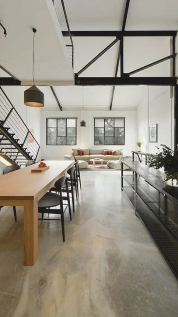 An open-plan luxury villa interior showcasing a large expanse of seamless polished concrete flooring. The light sand-grey concrete floor features a natural, matte architectural finish that flows smoothly from a foreground dining space into a sunlit living room. The industrial-modern architecture features a high vaulted ceiling with exposed matte black structural steel trusses, a matching steel-frame open staircase, and clean white walls, showing how a monolithic concrete base anchors a bright and airy upscale home.