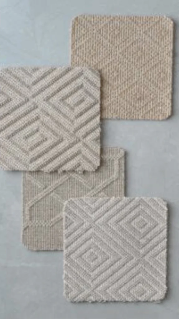 A top-down view of four square carpet samples made of natural fibers, arranged overlapping on a smooth, grey stone surface. Each sample showcases a high-quality, tightly woven neutral tone—ranging from warm beige and soft tan to cool grey. The textiles feature a distinct, geometric diamond and ribbed pattern woven directly into the fabric, providing a highly textured, luxurious feel designed for layering and adding soft architectural depth to a high-end space.