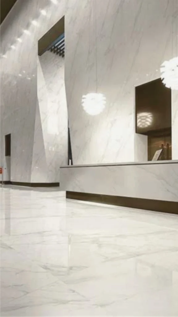 A striking open-concept luxury villa living space featuring a vast expanse of large-format grey porcelain flooring that mimics natural marble. The large slabs have ultra-fine, color-matched grout lines, giving the floor a completely seamless, continuous appearance. The polished stone look reflects natural light beautifully, complementing a sleek white fabric sectional sofa and a dark wood coffee table. In the background, a massive matching marble or porcelain feature wall stands adjacent to floor-to-ceiling windows, exemplifying high-end, monolithic architectural design.