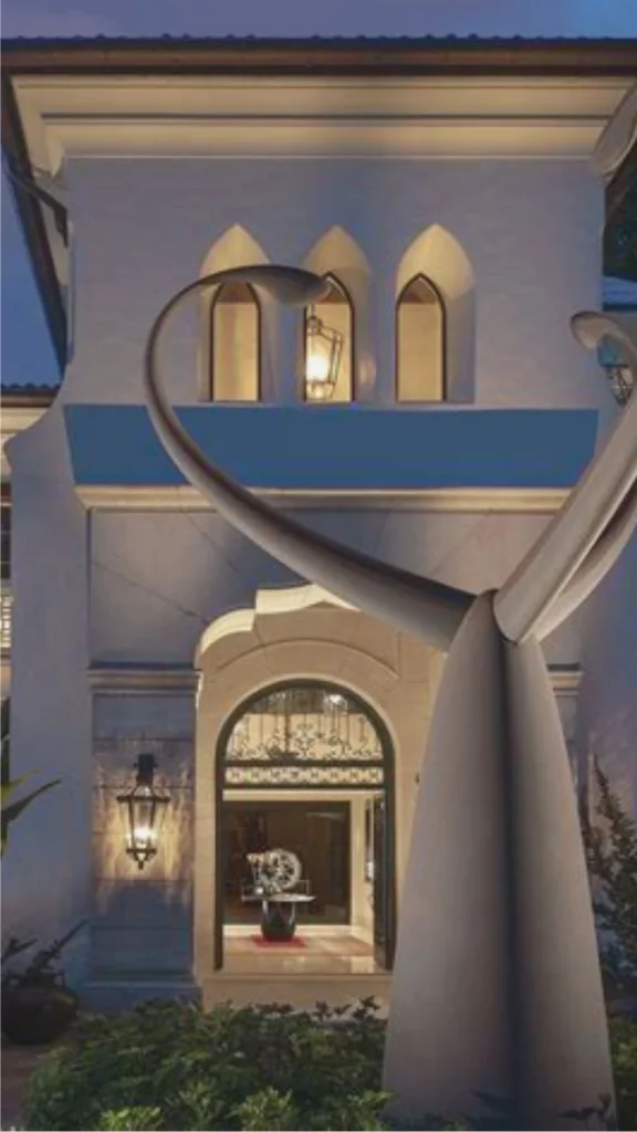 An elegant exterior entryway of a high-end estate showcasing sophisticated integrated lighting and luxury villa design. The white textured stucco facade features architectural details like an arched entry portal and a second-story balcony with three pointed arch windows, softly illuminated by concealed warm lighting. A classic black iron lantern glows on the stone wall beside the entrance, while a large, modern curved metal sculpture in the foreground stands dramatically lit, emphasizing texture and depth without any visible bulbs or visual clutter.