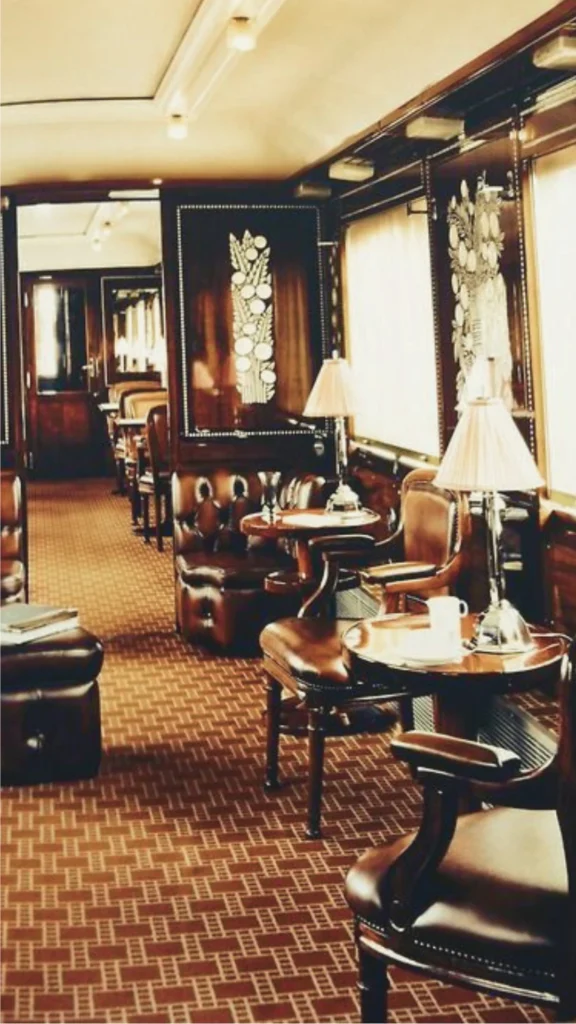 A detailed view of a premium luxury train cabin interior showcasing high-end, stable materials designed for travel on rails. The foreground features a small dining table made of rich, dark solid hardwood, beautifully set with fine porcelain dishware and polished glassware. Along the wall, comfortable seating is upholstered in a clean, tight-woven neutral fabric with decorative bolster pillows. A large window frames a passing scenic landscape, while warm, hidden accent lighting reflects off the smooth wood wall paneling and polished brass fixtures, demonstrating a quiet and sophisticated layout.