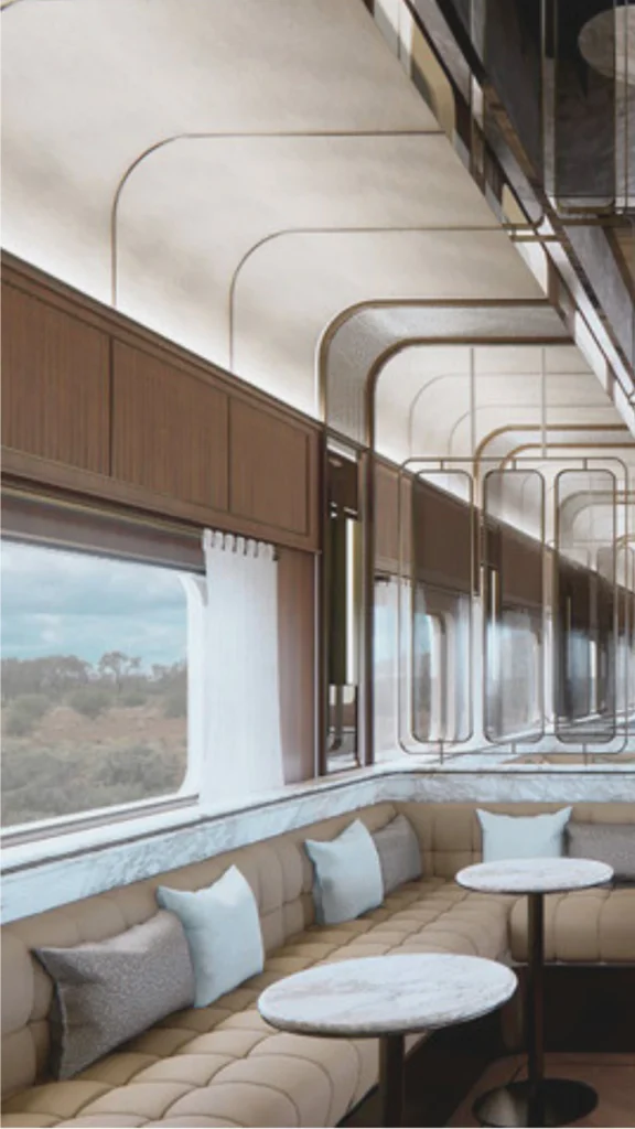 A detailed view of a premium luxury train cabin interior showcasing high-end, stable materials designed for travel on rails. The foreground features a small dining table made of rich, dark solid hardwood, beautifully set with fine porcelain dishware and polished glassware. Along the wall, comfortable seating is upholstered in a clean, tight-woven neutral fabric with decorative bolster pillows. A large window frames a passing scenic landscape, while warm, hidden accent lighting reflects off the smooth wood wall paneling and polished brass fixtures, demonstrating a quiet and sophisticated layout.
