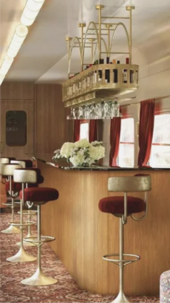 A sleek, modern luxury train lounge car highlighting an advanced layered lighting system. Long, continuous rows of warm white ambient LED strip lights are seamlessly recessed into the architectural ceiling coves, throwing a soft, indirect glow down across the cabin. Below, an expansive tufted tan leather banquette sofa runs parallel to a large panoramic window framing an outdoor landscape. Minimalist, round white marble cocktail tables break up the seating layout, while tinted glass privacy screens and polished metallic trim reflect the hidden, glare-free illumination to make the long car feel open and expansive.
