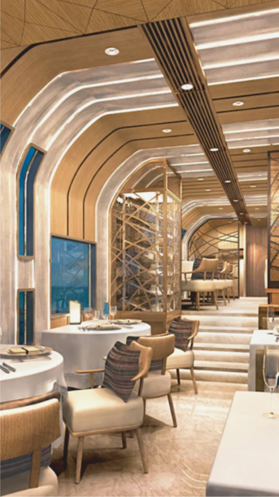 A sophisticated luxury train dining car showcasing a perfectly balanced warm neutral color palette. The cabin features curved wood-grained ceiling and wall panels in a light greige and caramel tone, seamlessly integrated with continuous rows of glowing white ambient lighting to make the space feel higher. Upscale dining tables dressed in crisp white linens are paired with plush cream-colored armchairs accented by textured, muted gold and deep brown striped pillows. In the background, low marble steps lead to an elevated seating section framed by an elegant, geometric gold mesh partition screen that coordinates with the highly polished, light stone flooring.