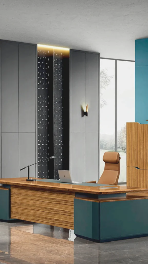 A striking example of modern executive office interiors luxury showcasing a sculptural and substantial workspace design. The focal point is a massive, premium wooden desk with rich grain textures, anchored by smooth, matte teal geometric base cabinets. An ergonomic high-backed executive chair upholstered in warm tan full-grain leather complements the desk layout. The background features floor-to-ceiling minimalist grey wall paneling accented by a narrow, dark textured vertical alcove with warm built-in backlit illumination and a modern gold sconce, delivering a high-performance workspace with instant professional gravitas.