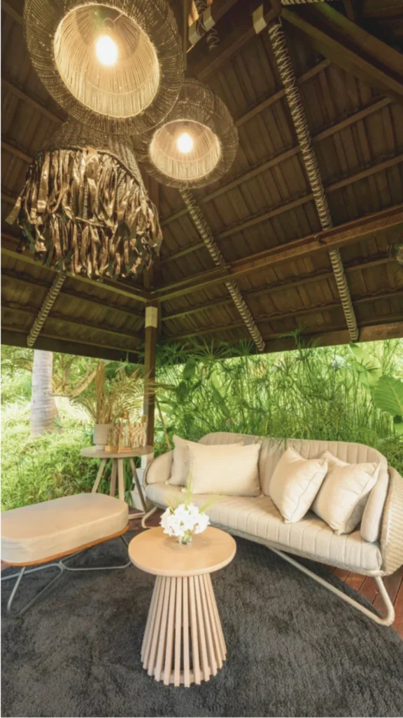 A luxurious, open-air nature retreat lounge set beneath a rustic wooden pavilion roof. The space features a chic beige sofa with plush throw pillows, a matching ottoman, and a round wooden slatted coffee table resting on a dark gray area rug. Massive woven rattan pendant lights hang from the exposed ceiling, casting a warm glow over the seating area. The pavilion is entirely open to the lush, vibrant green tropical foliage and tall grass surrounding it, effortlessly blurring the line between indoor luxury and outdoor nature.