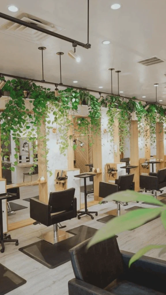 A sleek and modern hair salon interior featuring a row of styling stations. Each station is equipped with a black leather salon chair, a tall rectangular mirror framed by light wood pillars, and minimalist black utility carts. An abundant, vibrant canopy of green leafy vines cascades down from the top shelf of the wooden pillars over the mirrors, adding a refreshing, natural biophilic element to the bright space. The salon has light wood flooring, a clean white ceiling with recessed lighting, and industrial black track lights focused on each station.