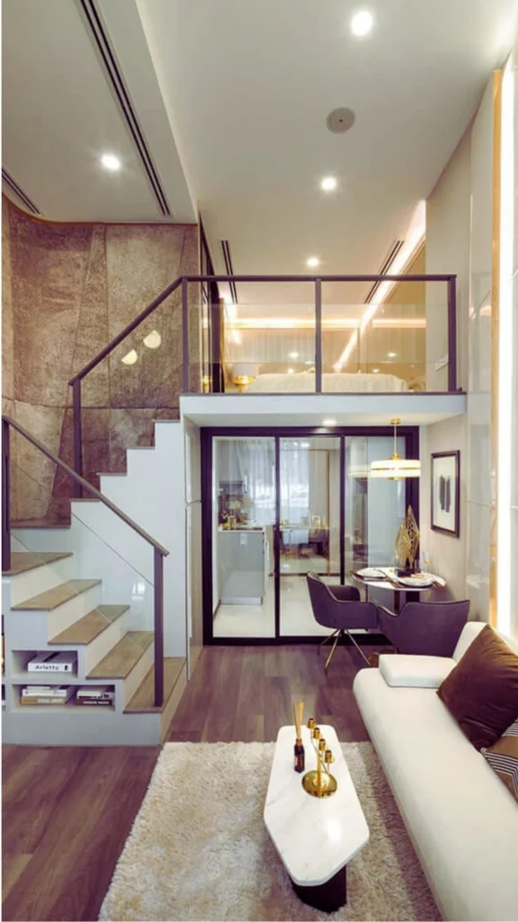 A wide shot of a modern, multi-level luxury loft interior from the image, highlighting an upscale condo design with a calm, neutral palette. In the foreground, a sleek cream-colored sofa is styled with dark brown and geometric-patterned pillows, facing a minimalist white marble coffee table on a plush, high-pile beige area rug. To the left, a light grey staircase with a dark metal and glass railing features built-in display shelving underneath, leading up to a lofted bedroom level protected by a glass balustrade. The left wall boasts a dramatic textured accent finish in earthy brown tones, while the double-height right wall features glossy vertical panels integrated with glowing linear LED light strips. In the background, a small dining set with charcoal grey armchairs sits next to a glass sliding door that reveals a modern kitchen.