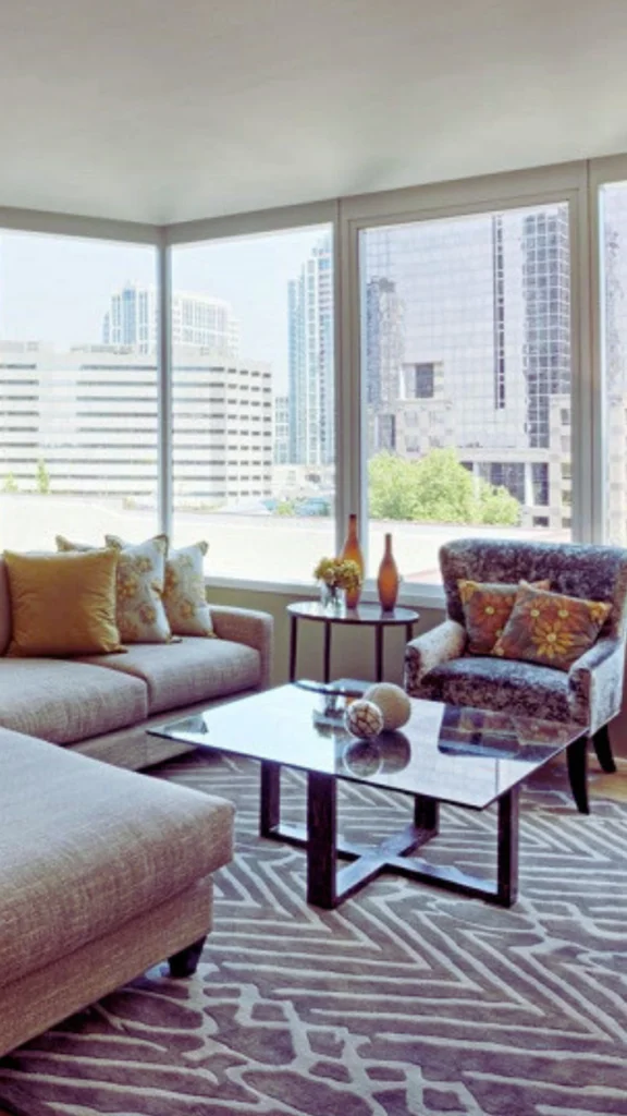 A modern high-end condo living room with large floor-to-ceiling windows showing a city skyline. The room features a neutral sofa, a patterned armchair, a geometric area rug, and a prominent dark wood coffee table with a glass top acting as the room's center piece.