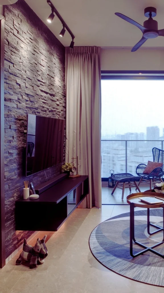 An upscale condo living room showcasing a rich mix of materials, featuring a prominent textured dark gray stone accent wall with a wall-mounted TV. The space balances stone texture with smooth flooring, a dark wooden media console, a metal-framed coffee table, and an adjacent balcony with woven patio seating.