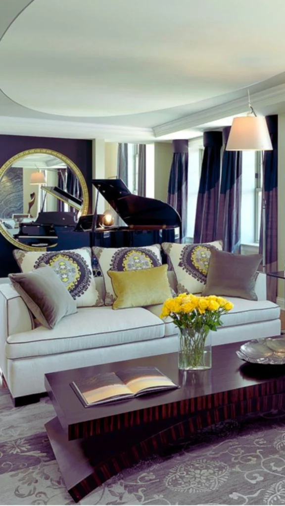 An upscale condo living room utilizing smart reflection to expand the space. A large, round mirror with a gold frame is mounted on a dark accent wall behind a white sofa, reflecting a black grand piano, windows with deep purple drapes, and open sightlines across the room.