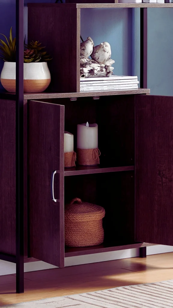 A close-up of a modern dark wood storage cabinet with its doors swung open, showcasing organized internal shelving. Inside, a small woven basket and wrapped decorative candles are neatly hidden away, while the upper open shelving features intentionally styled items, including a stack of magazines, a small bird sculpture, and a potted succulent.