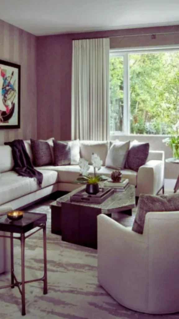 An elegant upscale condo living room featuring a framed abstract artwork mounted on a muted purple accent wall. The space is softened with a large off-white sectional sofa, a white orchid centerpiece on a multi-tiered coffee table, and an accent chair, all positioned by a large window overlooking lush green trees.