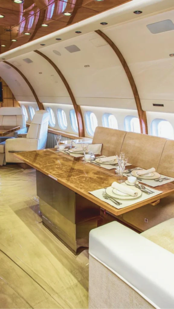 A luxurious VIP aircraft interior designed to look like a high-end dining room. It features a long, polished burl wood dining table set with fine china and glassware, a beige upholstered bench seat, and elegant wooden arches along the curved cabin ceiling, creating a residential-style layout on a private jet.