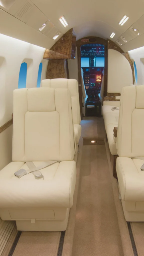 Interior view of a luxury private aircraft cabin looking down a beige carpeted center aisle toward the cockpit. The seating arrangement features oversized cream leather executive chairs and a side-facing leather sofa bench. The cabin is lit with bright overhead panel lights and accented with polished burl wood partition